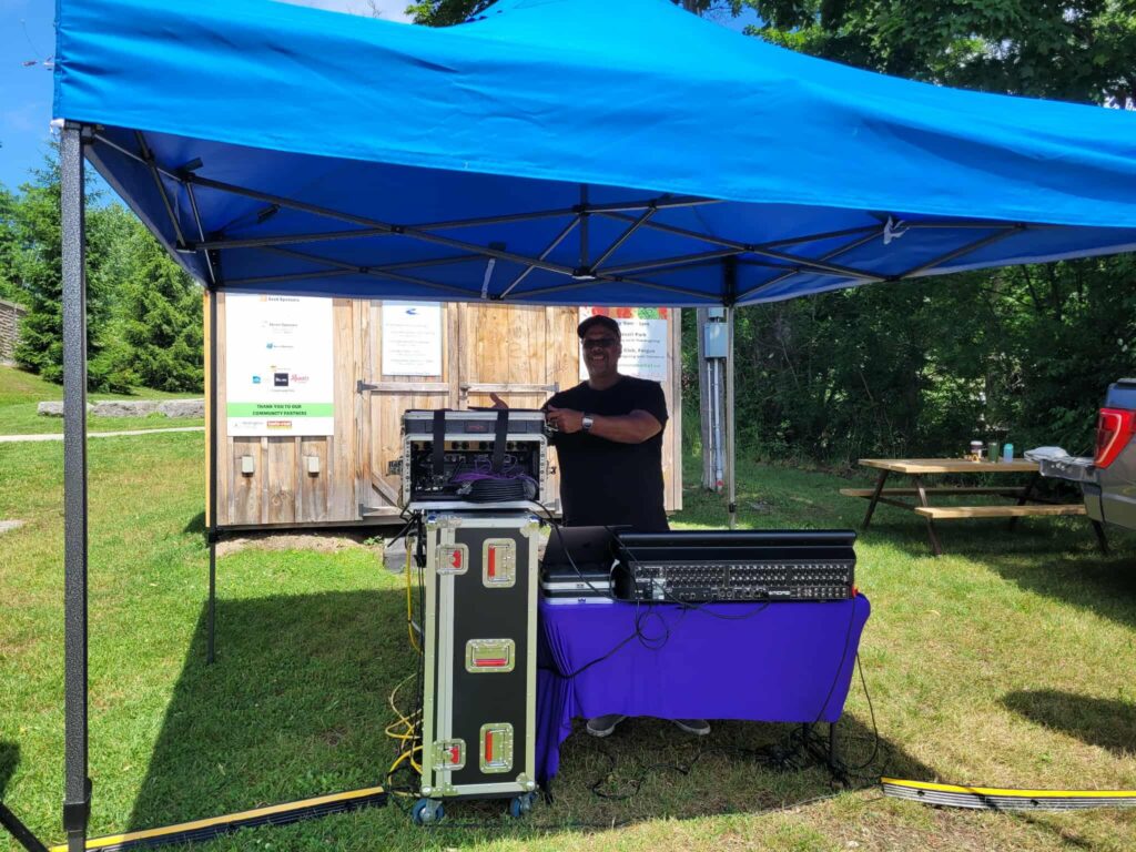 A DJ and sound mixing setup under a blue canopy, with gear and a purple cloth table at a daytime Canada Day event in a small town park.