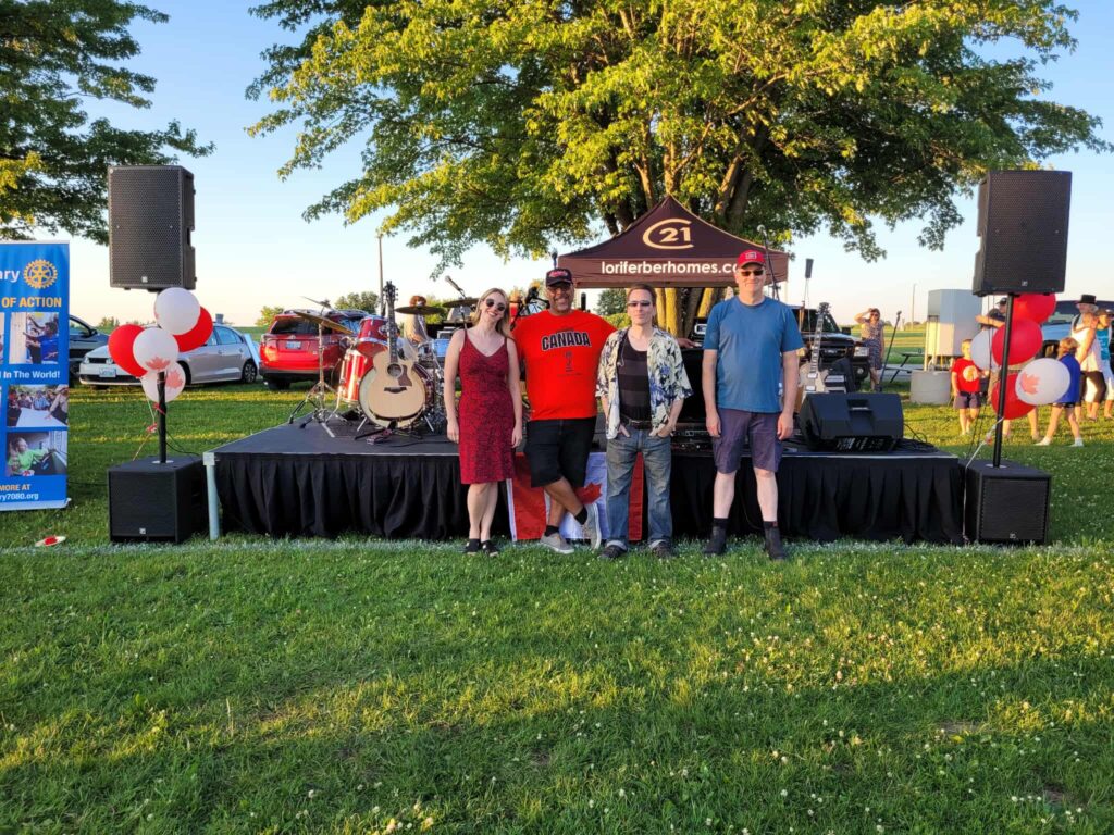 Four members of a Canadian band posing in front of their instruments and stage setup at sunset during Canada Day festivities.