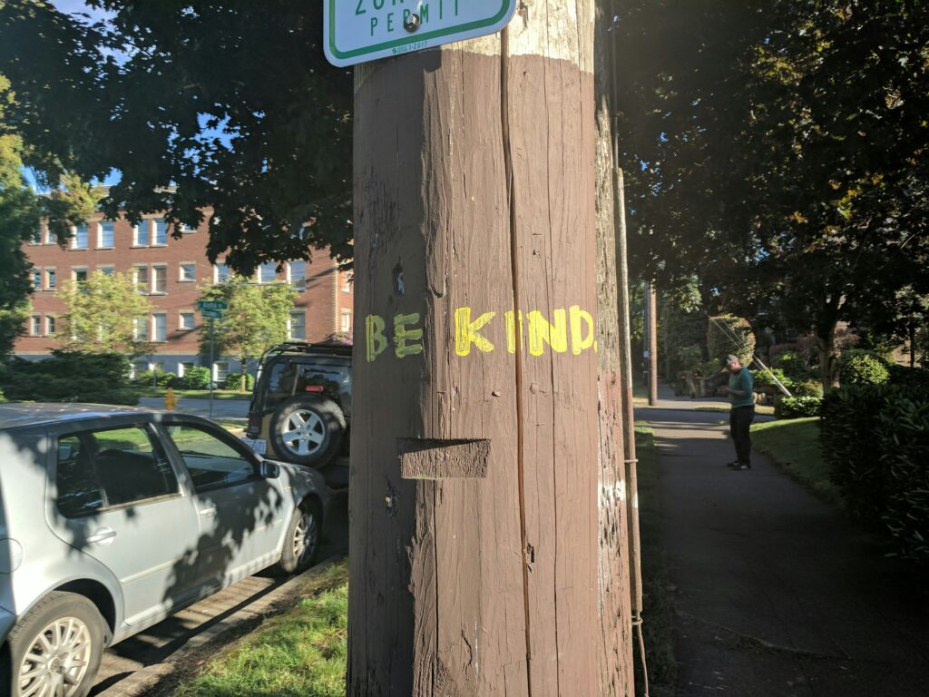 bright street with 'be kind' written in yellow paint on a hydro pole. 
